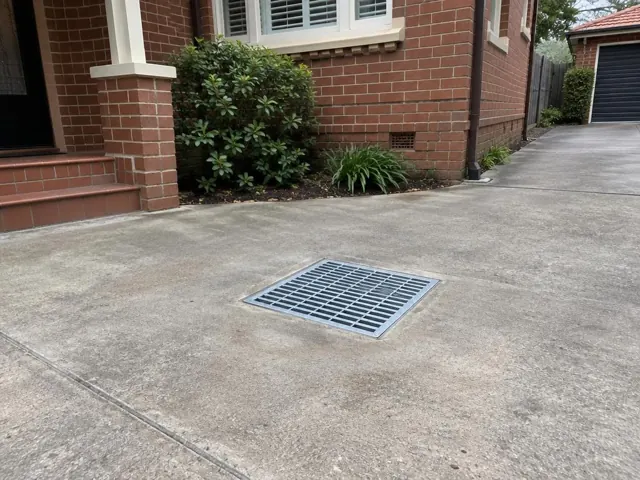 Clean square drain grate set in a concrete driveway beside a brick house in Naremburn